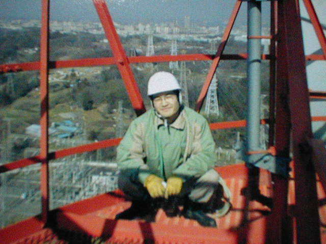 高層タワーの赤の鉄骨の上で、白いヘルメットと作業着姿の松原先生